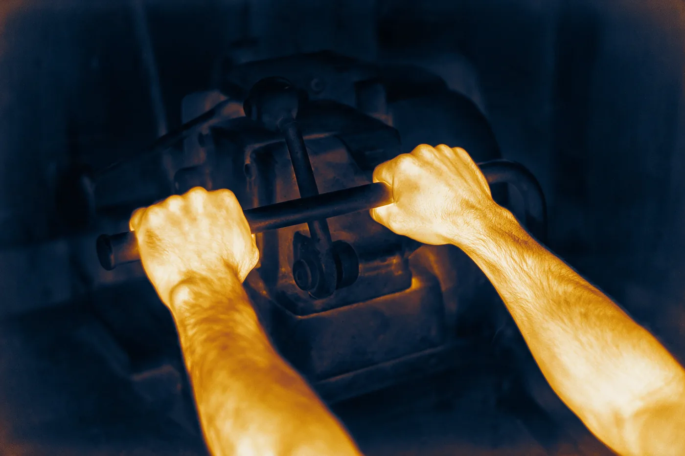 Lectura térmica de los antebrazos y las manos de un operario accionando la palanca de una máquina: la piel aparece en tonos dorados por el calor y la máquina en azul profundo.